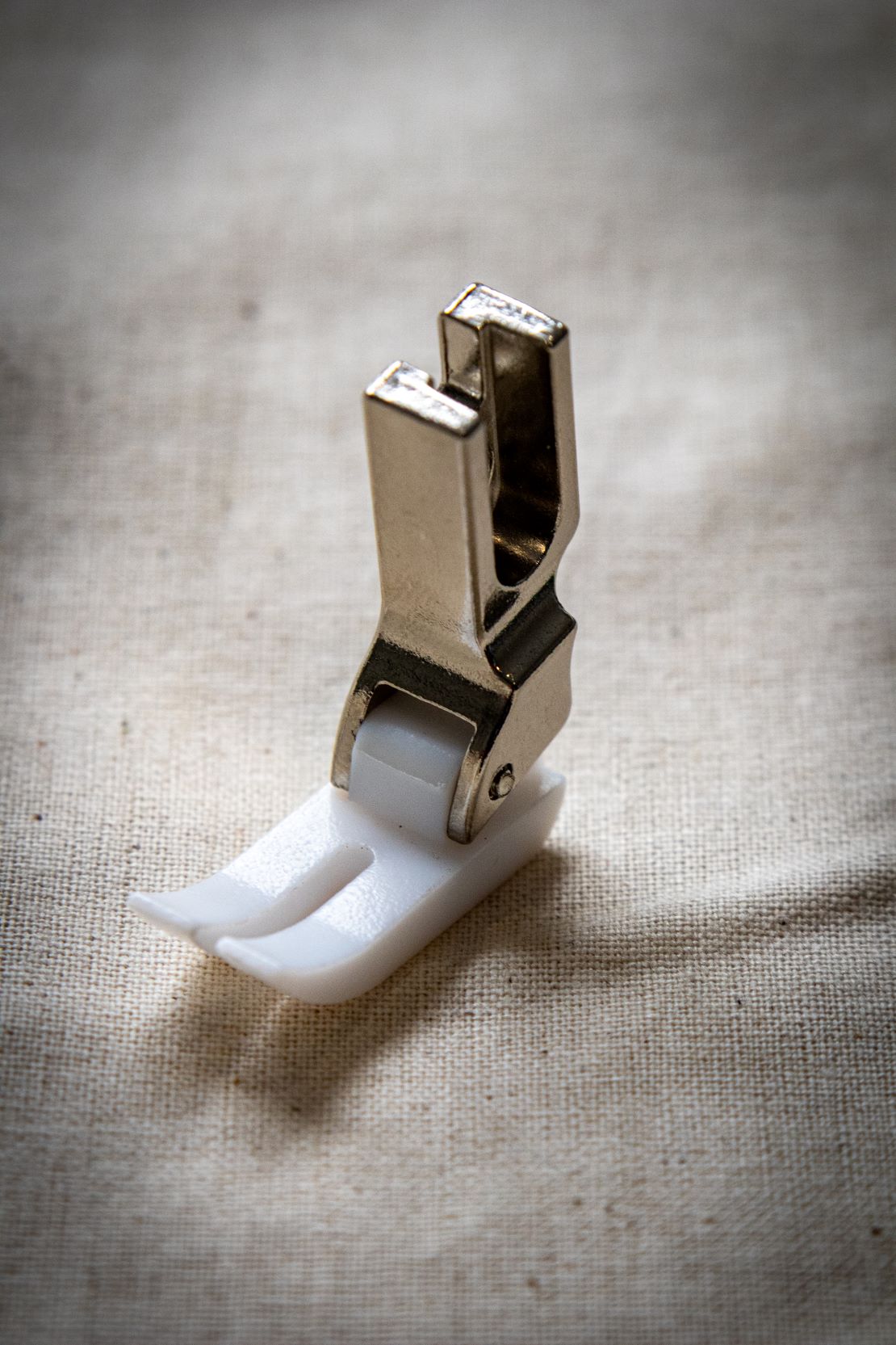 Angled view of the Teflon presser foot from the RMIT Store, showing a clear plastic upper section and white non-stick base, positioned upright on a beige fabric background.
